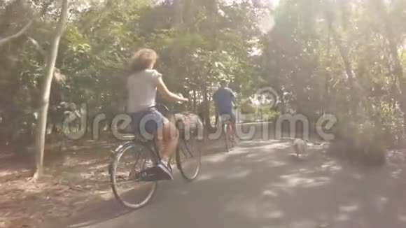 在公园里骑自行车的一对年轻快乐的嬉皮士夫妇和宠物狗在附近奔跑高清慢动作视频的预览图