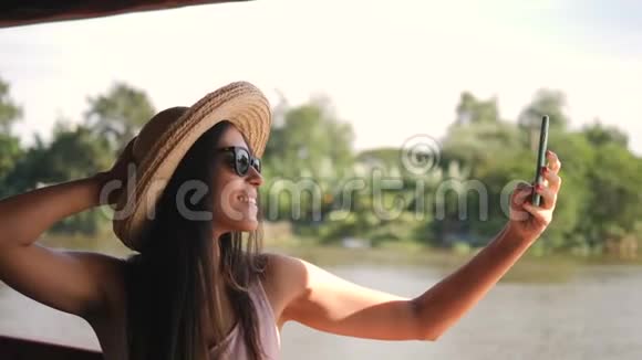 大帽子里美丽的混血旅游女孩在河边用手机和游船拍照视频的预览图