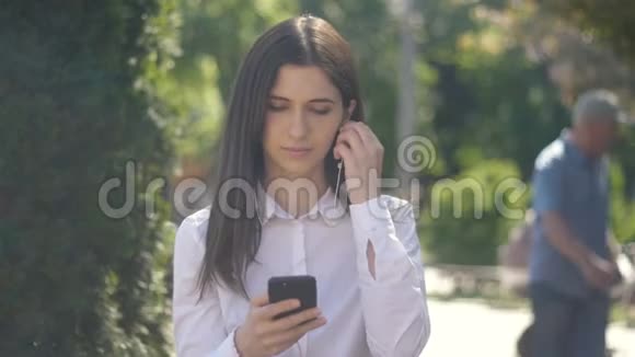 侧面观看白色衬衫年轻女子的肖像使用耳机和智能手机听音乐城市室外视频的预览图