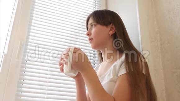 漂亮的女孩带着一杯热茶坐在窗台上微笑着视频的预览图