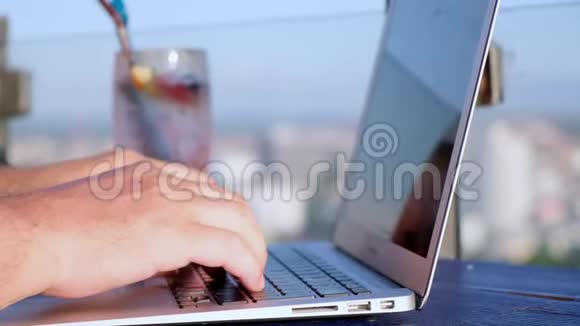 在一栋高楼的屋顶上的咖啡馆里男人在笔记本电脑上打字那里可以看到城市的美丽全景视频的预览图