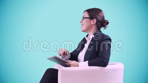 电视演播室戴眼镜的深色头发她穿着西装坐在工作室里接受采访蓝色视频的预览图