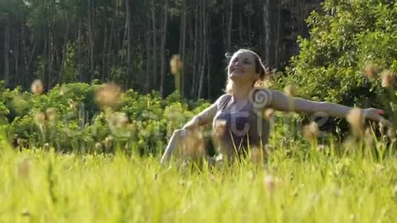 欢乐的女人坐在绿色的草坪上在夕阳的背景下快乐地举起双手飞向天空视频的预览图