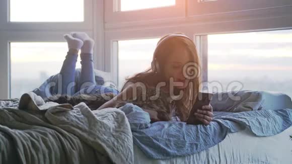 戴着耳机的年轻美女和她可爱的缅因州浣熊猫一起放松在窗边躺在床上听音乐视频的预览图
