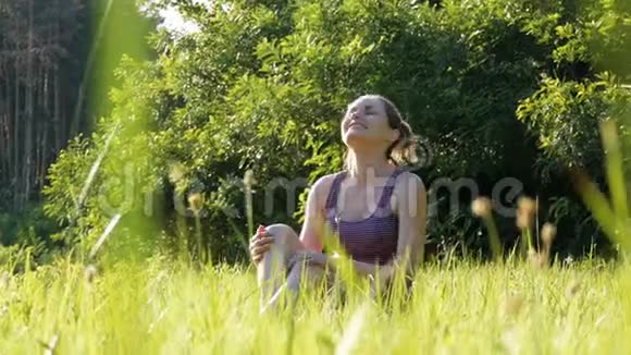 欢乐的女人坐在绿色的草坪上在夕阳的背景下快乐地举起双手飞向天空视频的预览图