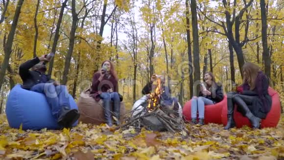 深色皮肤和白色的男人三个女人坐在秋天森林里的火炉旁用热水瓶慢慢地喝视频的预览图