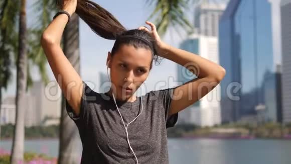 年轻的混合种族健身女子在晨跑前整理她的头发在城市公园和大型办公大楼视频的预览图