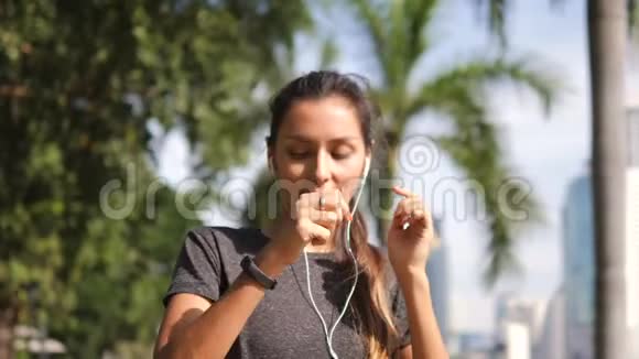 吸引人的年轻混合种族健身跑步者女士在城市公园的移动智能手机上打开耳机和打开音乐视频的预览图