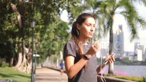 年轻的亚洲跑步女孩在城市公园的移动智能手机上打开耳机并打开音乐4K慢动作视频的预览图