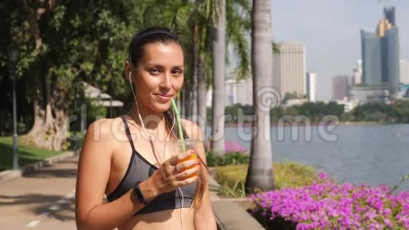 年轻的混合跑跑女孩喝新鲜橙汁和看镜头在城市公园4K慢动作泰国视频的预览图