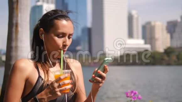 在城市公园与大城市公园晨跑后年轻跑步女子在手机上发短信喝新鲜橙汁视频的预览图