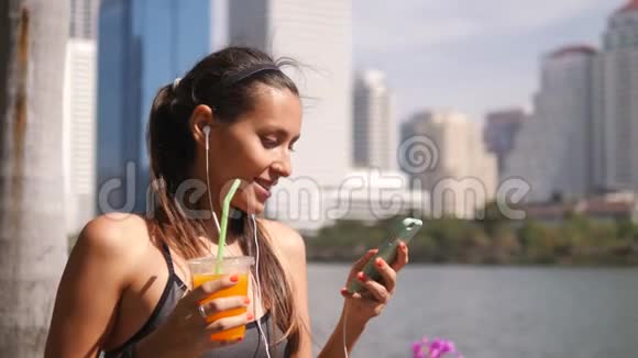吸引人的健身妇女使用移动应用程序和饮用新鲜橙汁后上午跑在城市公园与大视频的预览图