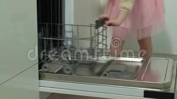 可爱的金发女孩穿着粉红色的裙子在明亮的厨房帮助妈妈在洗碗机里装盘子杯子视频的预览图