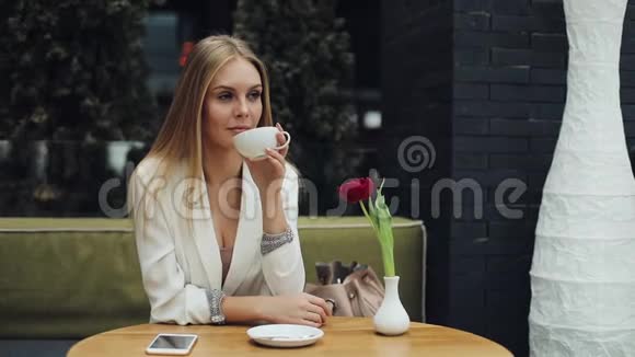一位年轻的金发女人端着一杯咖啡到她的嘴唇坐在咖啡馆的桌子旁视频的预览图