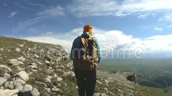 像个从后面戴帽子的女孩一位背着背包的游客沿着岩石高原走到悬崖边视频的预览图