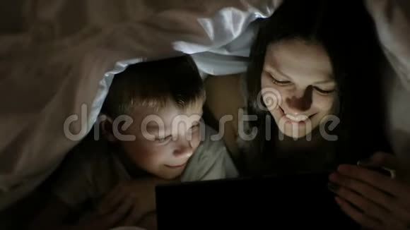 年轻女子妈妈和她的儿子一起在平板电脑上看有趣的电影在毯子下面笑视频的预览图