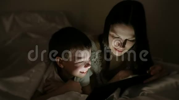 年轻女子妈妈和儿子一起在毯子下面的平板电脑上看一部有趣的电影视频的预览图