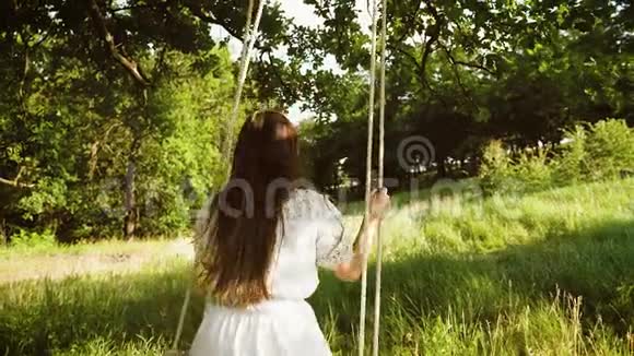 一个留着长发的女孩穿着白色连衣裙在夏天的公园里荡秋千慢动作视频的预览图