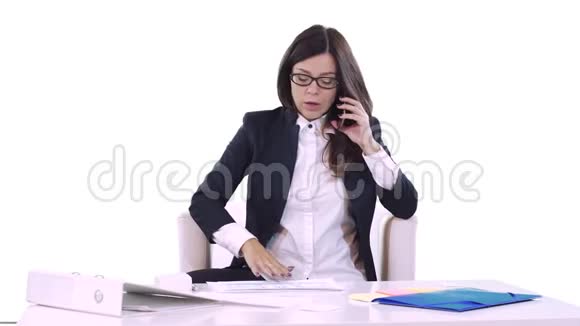 年轻的黑发女人坐在办公室的桌子上打电话桌子上有文件和视频的预览图