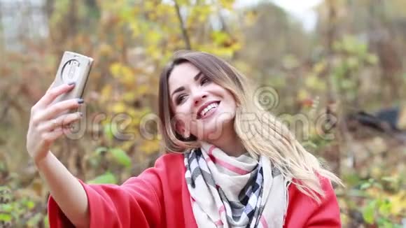 穿着红色外套和围巾的年轻女子带着手机在秋天的公园里视频的预览图