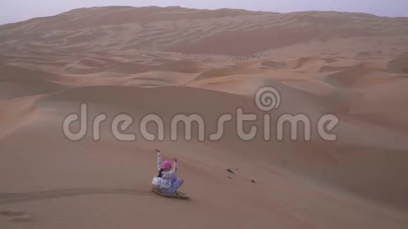 在阿拉伯联合酋长国的RubalKhali沙漠中的沙丘坡上少女在沙板上翻滚视频的预览图