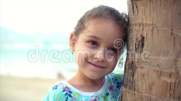 海滩上快乐的小女孩穿着一件蓝色的连衣裙戴着一棵棕榈树看着相机微笑着有点害羞儿童视频的预览图