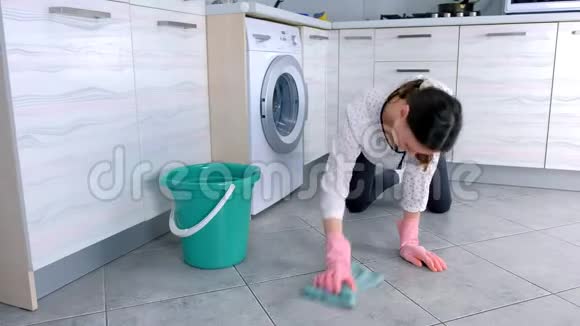 穿着粉红色橡胶手套的疲惫的女人用一块布洗厨房的地板地板上的灰色瓷砖视频的预览图