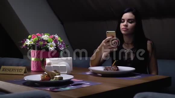 一位女士坐在靠近花束的餐馆里的一张预定的桌子旁在智能手机上拍摄智能菜肴的照片视频的预览图