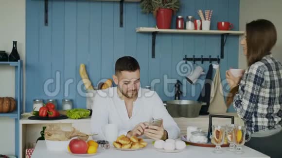 有魅力的一对夫妇在早餐时间在厨房见面并在家里使用智能手机进行社交媒体冲浪视频的预览图