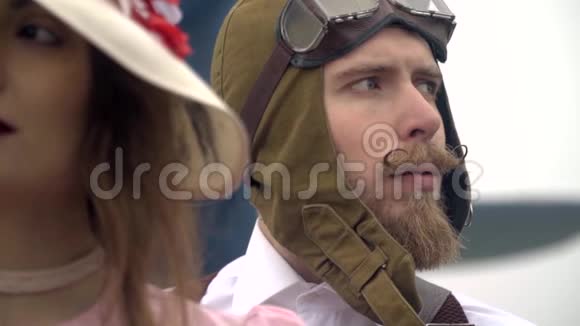 一个戴着头盔的留胡子的飞行员站在一个戴着第60号白帽子的女人旁边重新聚焦视频的预览图