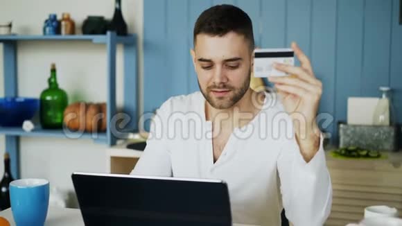 年轻人在家里的厨房里吃早餐时使用信用卡和笔记本电脑进行网上购物视频的预览图