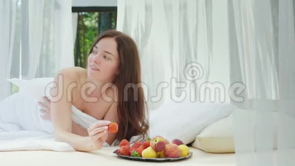 年轻的女人躺在床上手里拿着草莓在游泳池旁边的一个有窗帘的休息室里浪漫蜜月视频的预览图