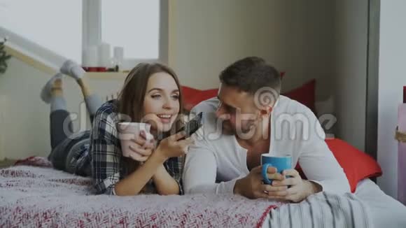 一对微笑的夫妇躺在床上看电视早上在家喝咖啡视频的预览图