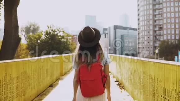 戴着长发的女孩在城市桥上散步穿着红色背包的黄墙之间的白人女人后视4K视频的预览图