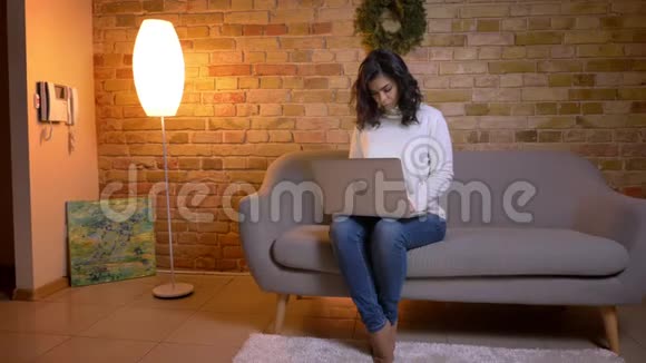 一幅专注的白种人黑发女商人坐在沙发上用笔记本电脑在家工作的肖像视频的预览图