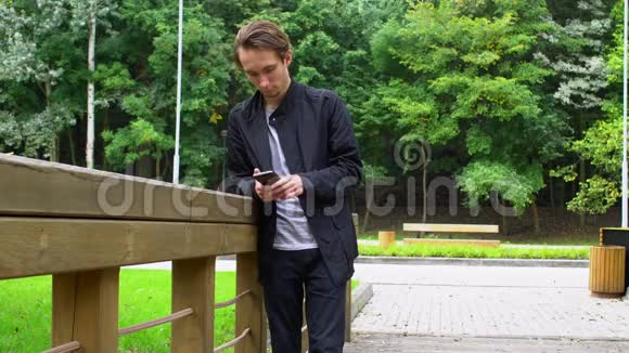 年轻人看着智能手机上的社交网络站在公园的桥上视频的预览图