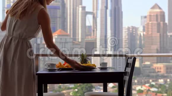 年轻的女家庭教师在阳台上准备早餐俯瞰市中心的摩天大楼视频的预览图