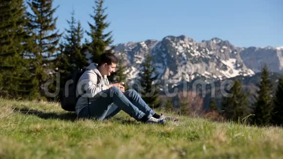 年轻的游客背着背包站在山顶上欣赏着令人惊叹的景色男人用智能手机拍照视频的预览图