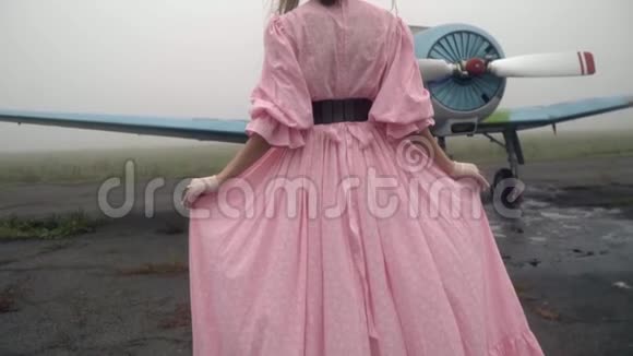 一个身材苗条的女人穿着一件粉红色的长裙走向站在老飞机机翼上的飞行员慢镜头视频的预览图