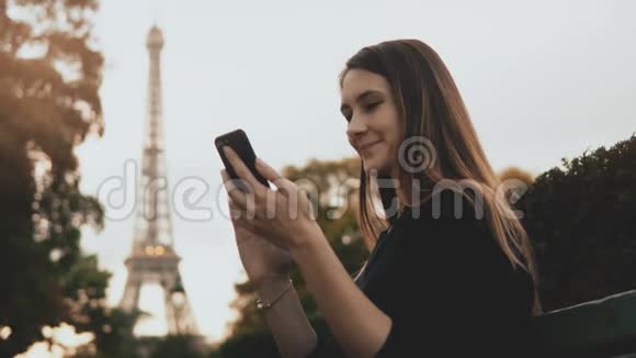 年轻漂亮的女人坐在法国巴黎埃菲尔铁塔附近的长凳上用智能手机视频的预览图