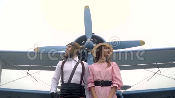 一个戴着头盔的野蛮飞行员站在一个穿着粉红色裙子的女人旁边她站在一架带螺旋桨的旧飞机旁视频的预览图
