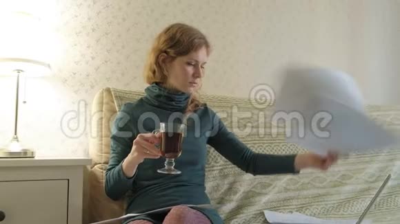 年轻女子穿着绿色连衣裙拿着笔记本电脑坐在沙发上用电话工作学习文件喝酒视频的预览图