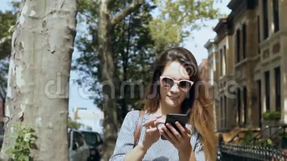 年轻漂亮的女人戴着太阳镜在阳光明媚的夏日走在外面用智能手机的肖像慢动作视频的预览图