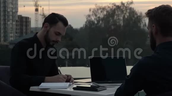 晚上两个商人带着笔记本电脑和电话在户外坐着一边讨论着什么一边微笑着视频的预览图