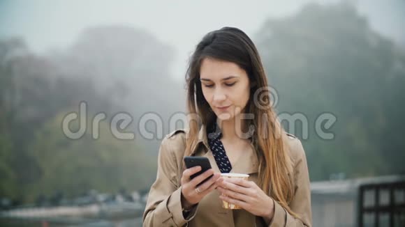 年轻美丽的女人在雾天清晨使用带有触摸屏的智能手机和喝咖啡的肖像视频的预览图