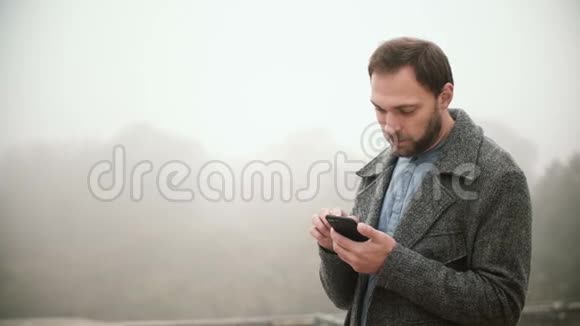 清晨站在雾蒙蒙的公园里的年轻英俊男子的肖像长胡子的男性使用带有触摸屏的智能手机视频的预览图
