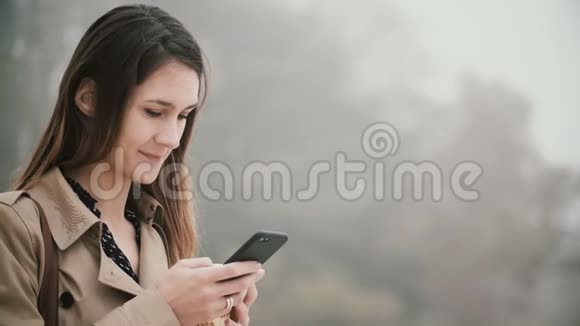 早晨雾蒙蒙的公园里年轻漂亮女人的肖像微笑的女性使用智能手机与触摸屏视频的预览图