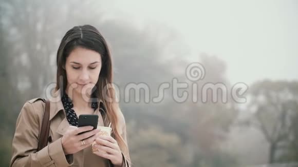 在雾蒙蒙的早晨年轻的美女站在公园里用智能手机喝着咖啡视频的预览图