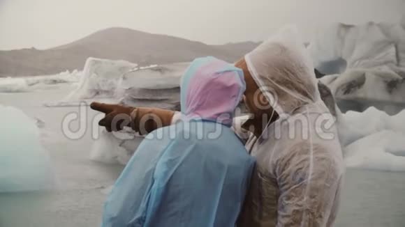 年轻夫妇穿着雨衣探索冰岛著名的景象冰泻湖游客男子向妇女展示一些东西视频的预览图