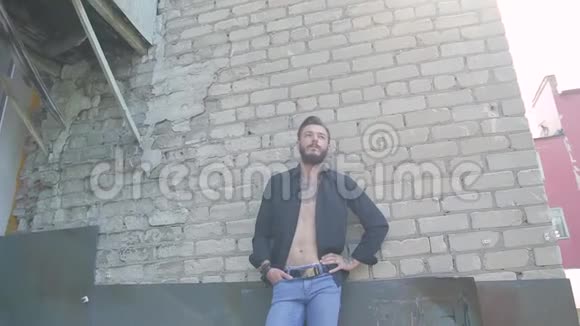 一个英俊的野蛮男人留着胡子穿着牛仔裤在灰色砖墙的背景上穿着一件黑色T恤视频的预览图